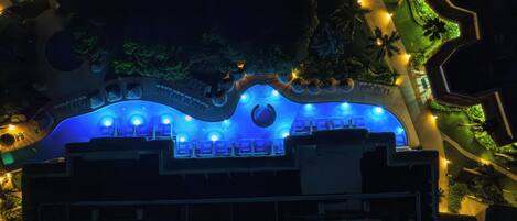 Vue aérienne d’un hôtel dont le toit présente un design original en forme de vague, illuminé de lumières bleues, et doté d’un jardin paysager avec des palmiers.