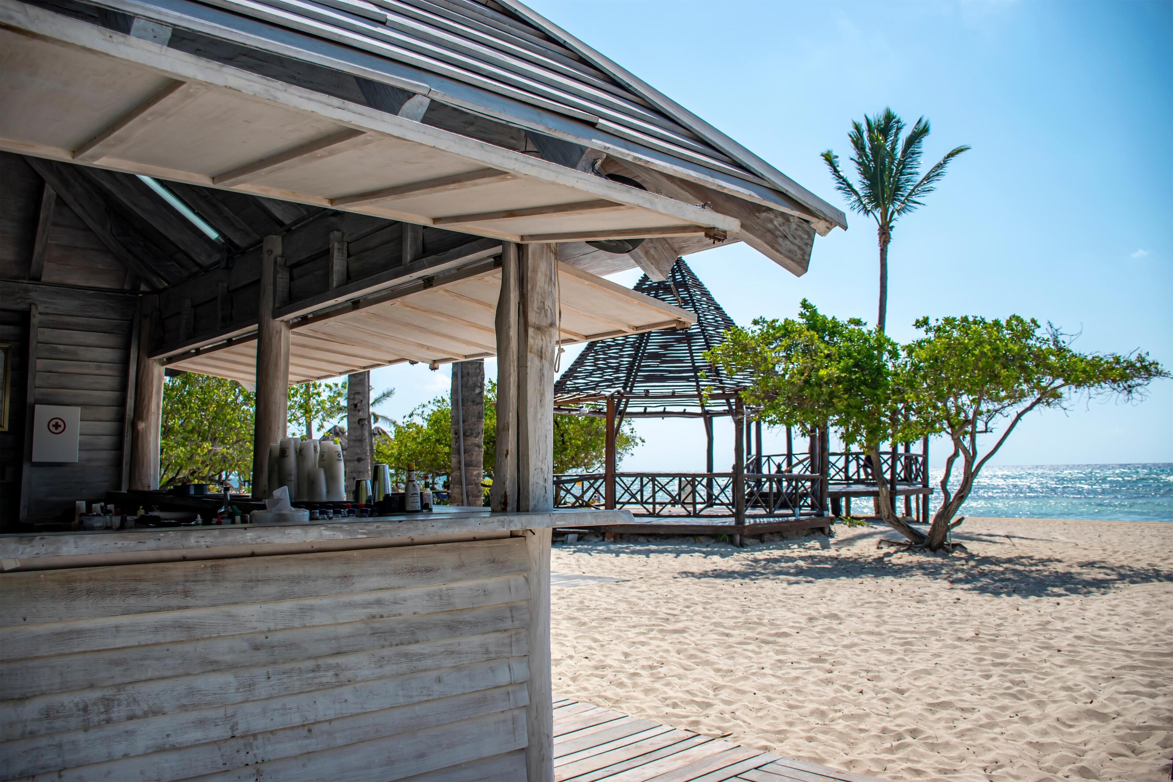 Una estructura de madera en la playa con un área de estar cubierta, una palmera y vista al océano.