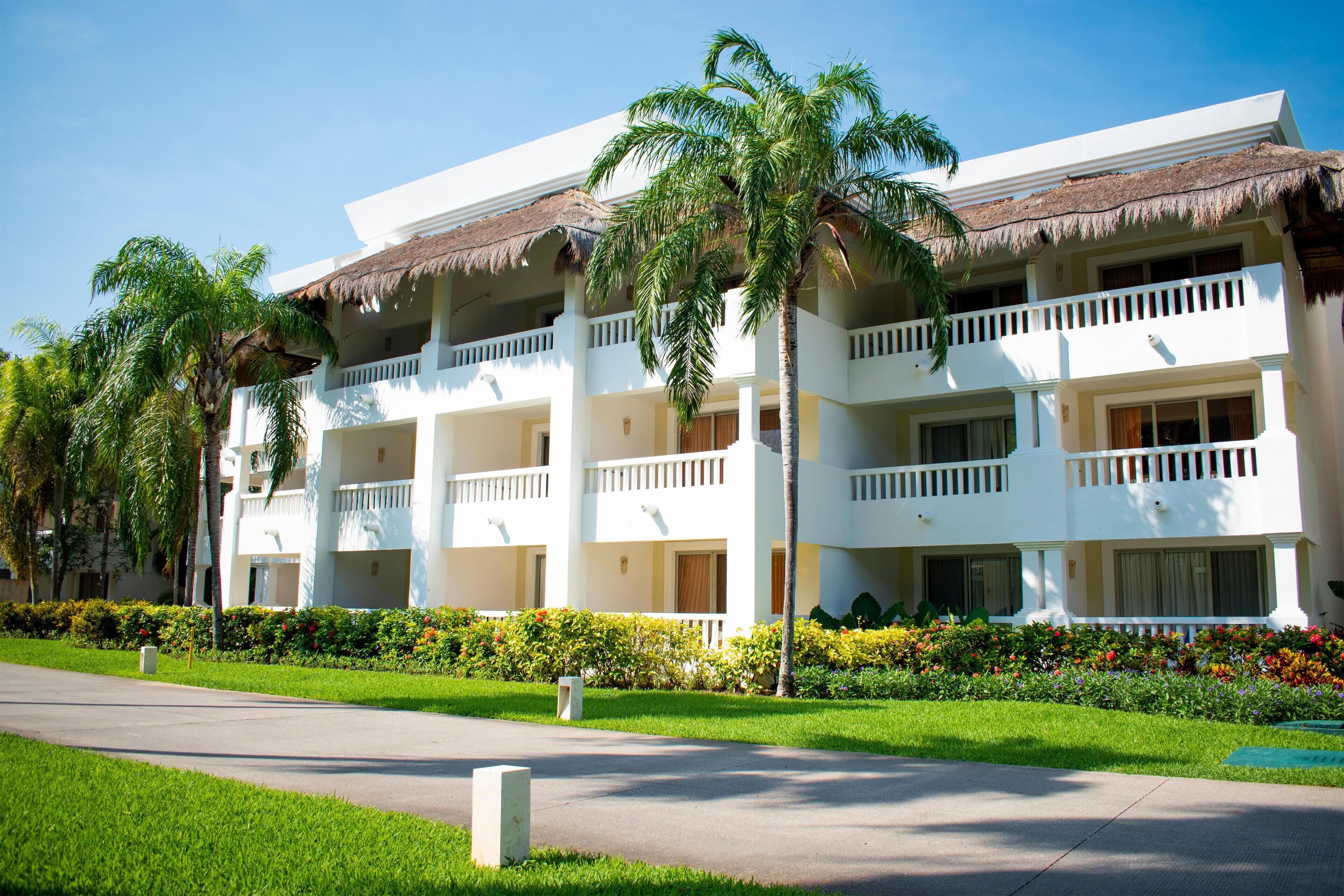 A multi-story hotel with a white facade, thatched roof balconies, and lush landscaping.