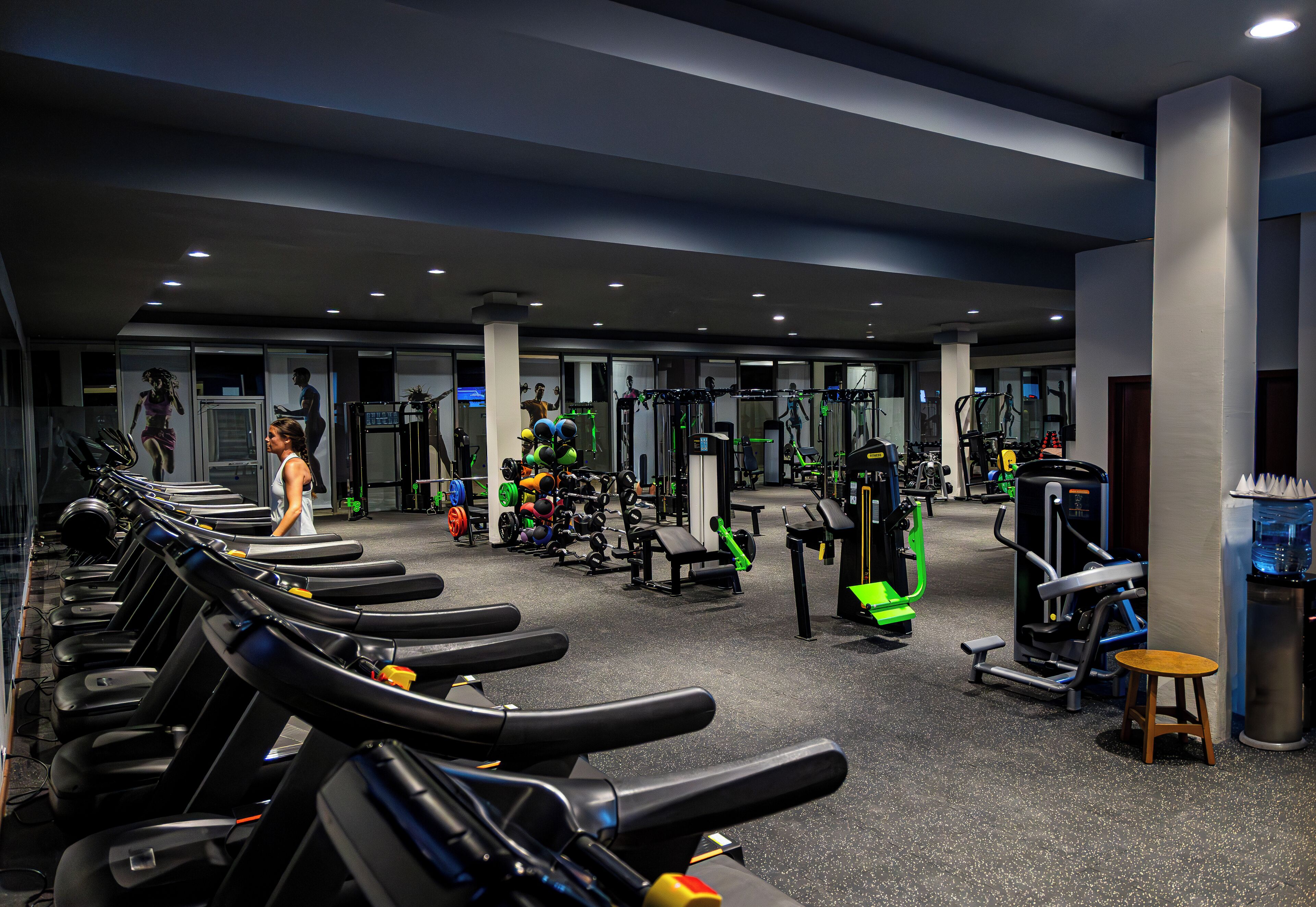 Une salle de sport équipée de tapis de course, d’appareils de musculation et d’un petit tabouret en bois.