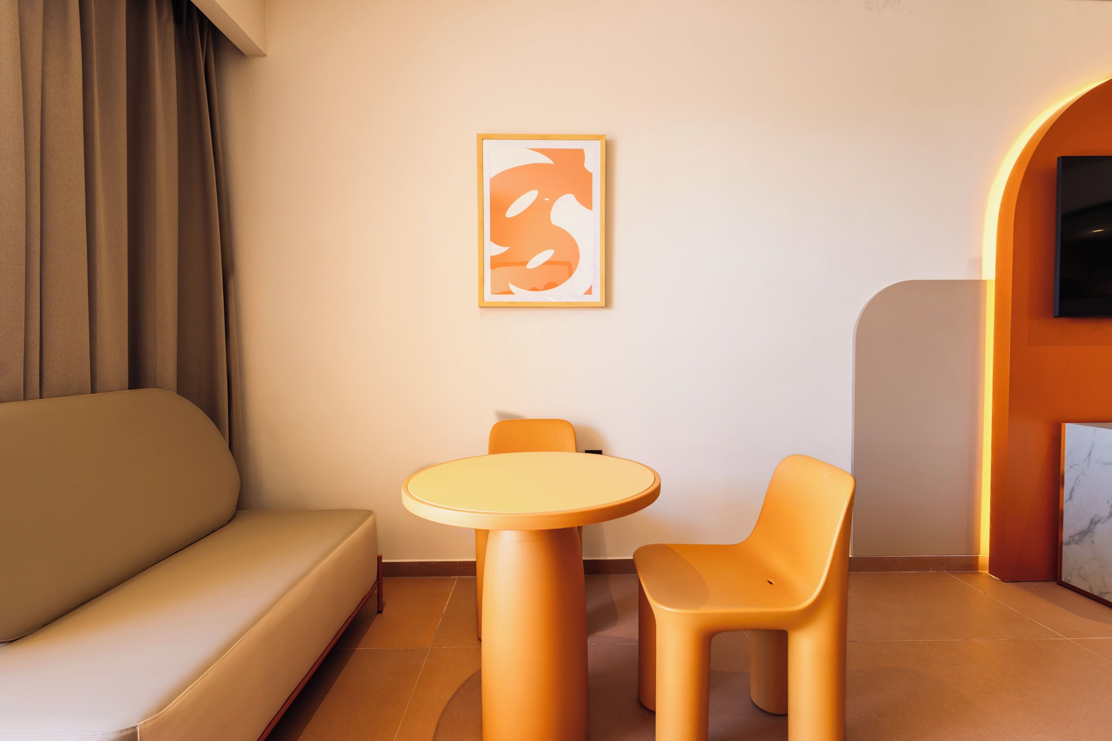 Una habitación de hotel moderna con un sofá, una mesa redonda, dos sillas, una obra de arte enmarcada y un televisor montado en la pared.