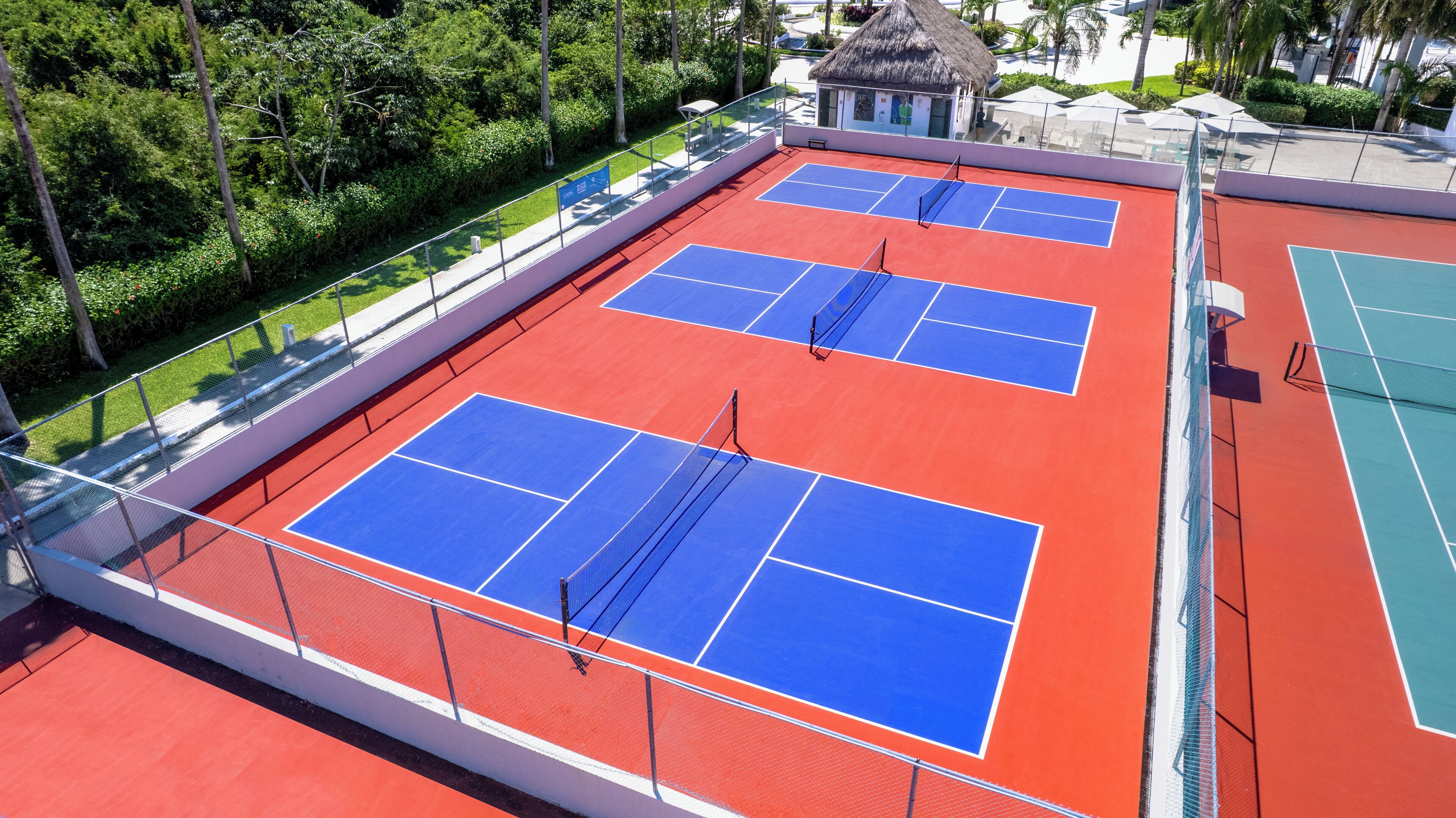 An outdoor tennis court with a red and blue surface, surrounded by a white fence and greenery.