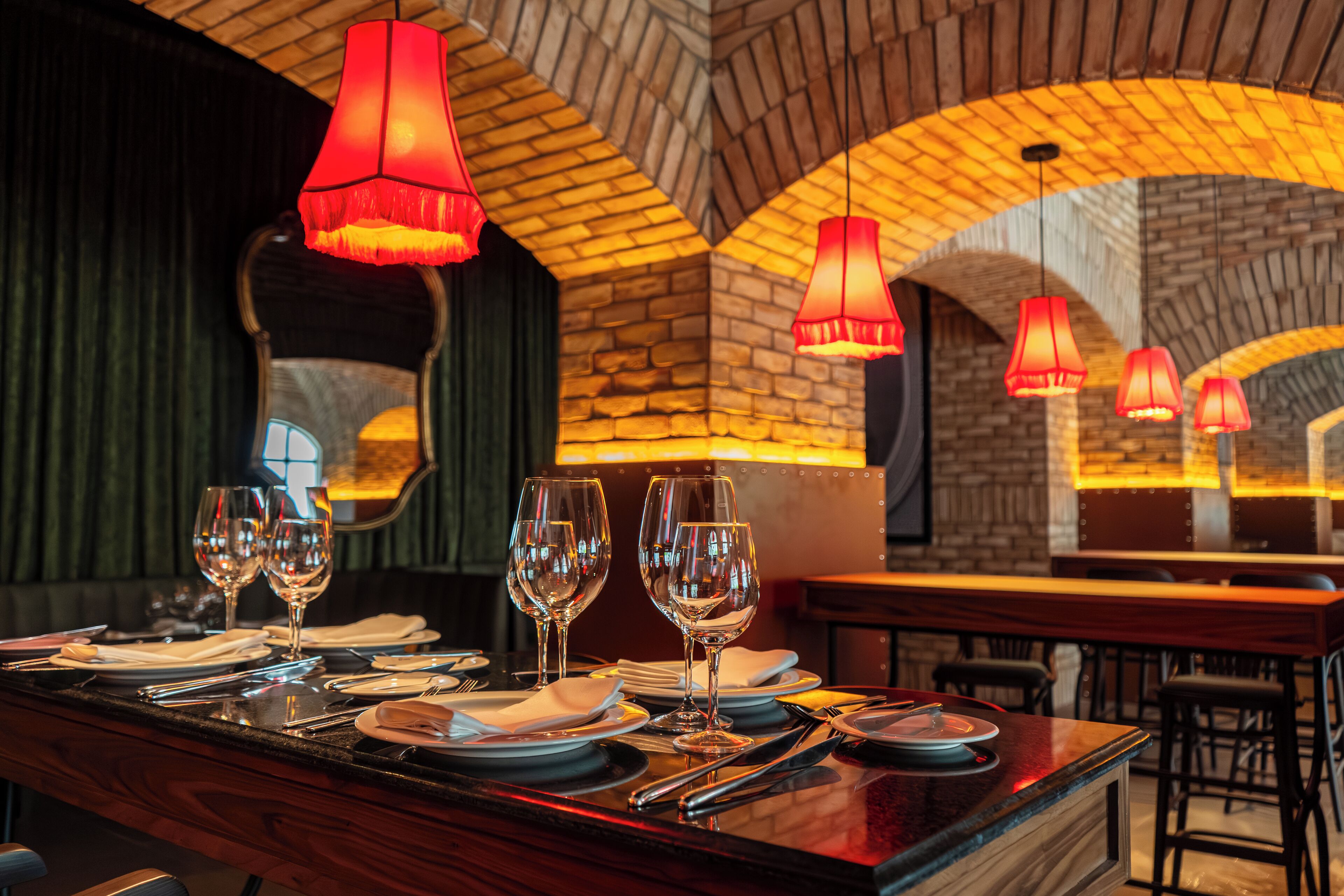 A restaurant with a brick wall and arched ceiling, featuring a bar area with neatly set tables and red pendant lighting.
