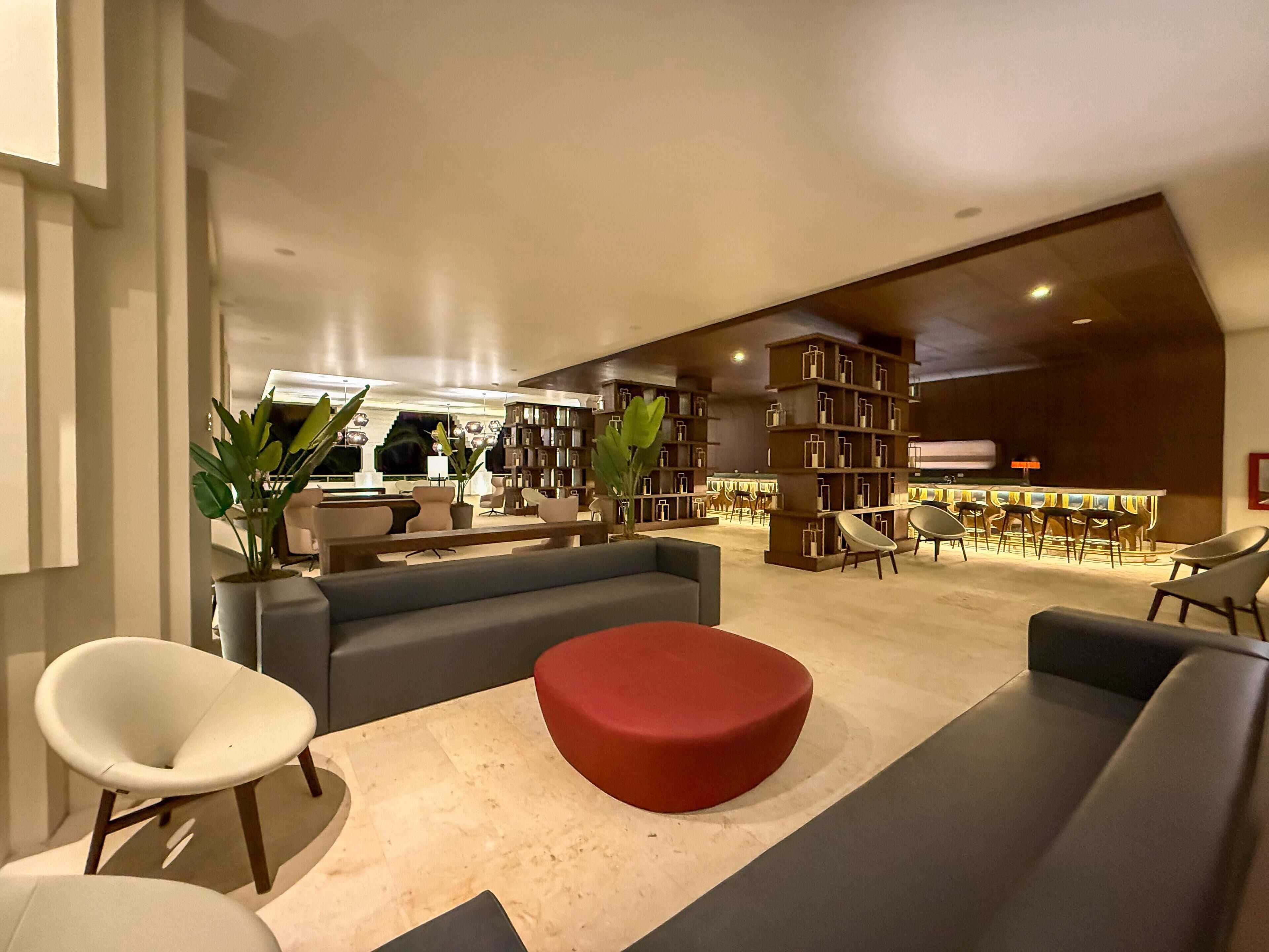A modern lounge area with sofas, a red ottoman, and a seating area with chairs. There are bookshelves filled with books and a bar area in the background.