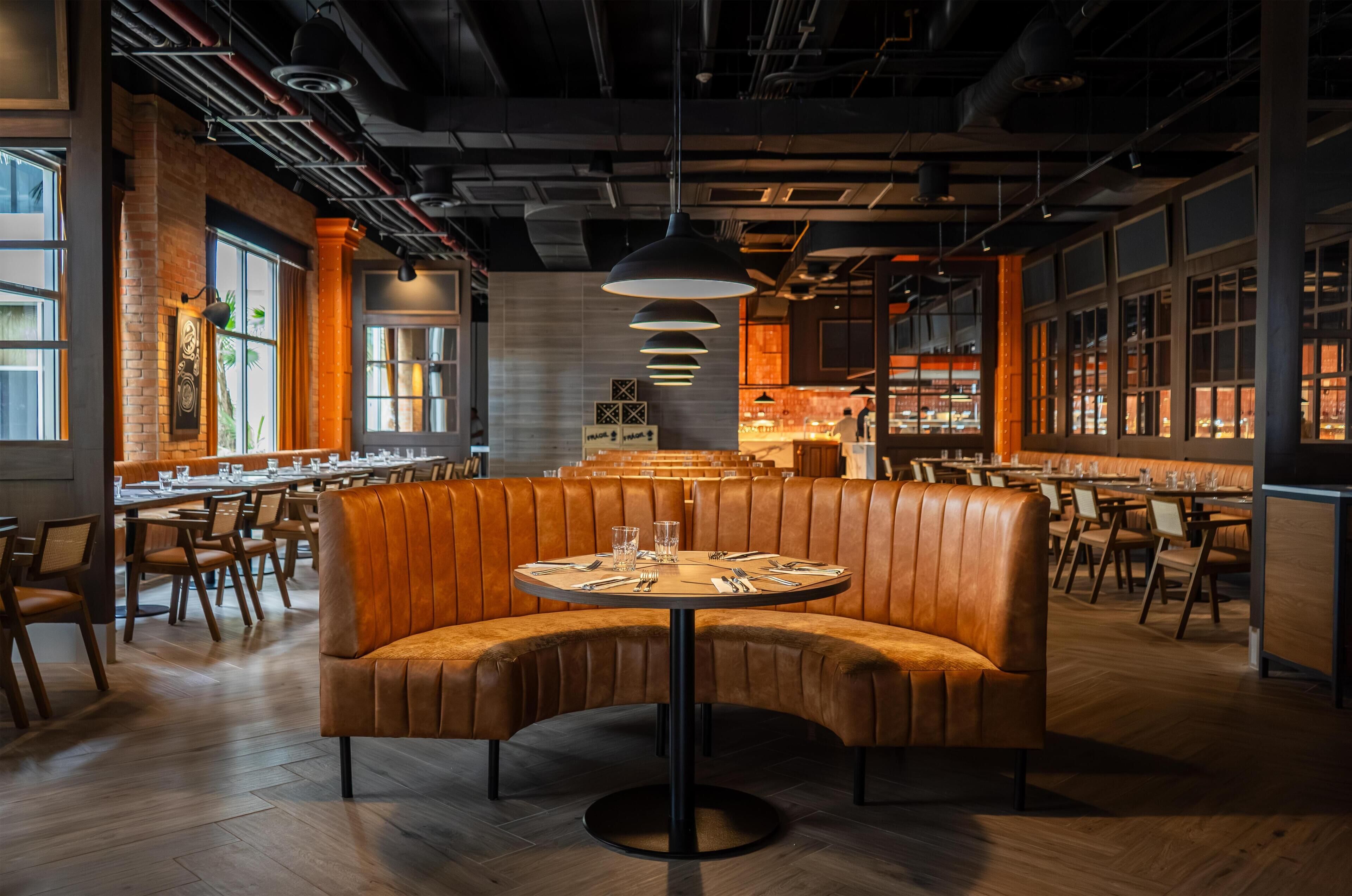 A restaurant with a central booth seating area, long communal tables, and a modern industrial ceiling design.