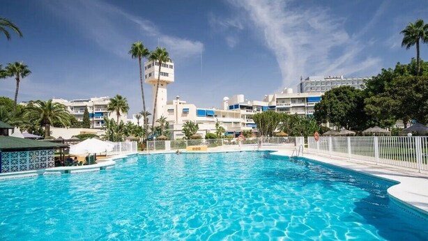 A swimming pool with a clear blue water, surrounded by a white fence, palm trees, and a tower-like structure in the background. - Beautiful Apartment Costa Lago on the Beach with Pool, Wi-Fi & Sea View; Parking Available (Torremolinos)