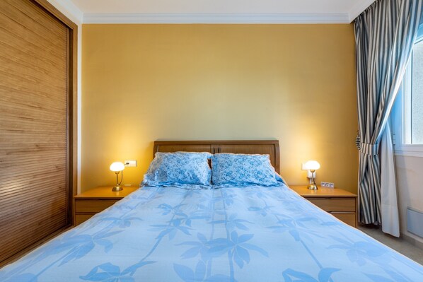 A hotel room with a bed, two bedside lamps, a wooden headboard, and a window with curtains.