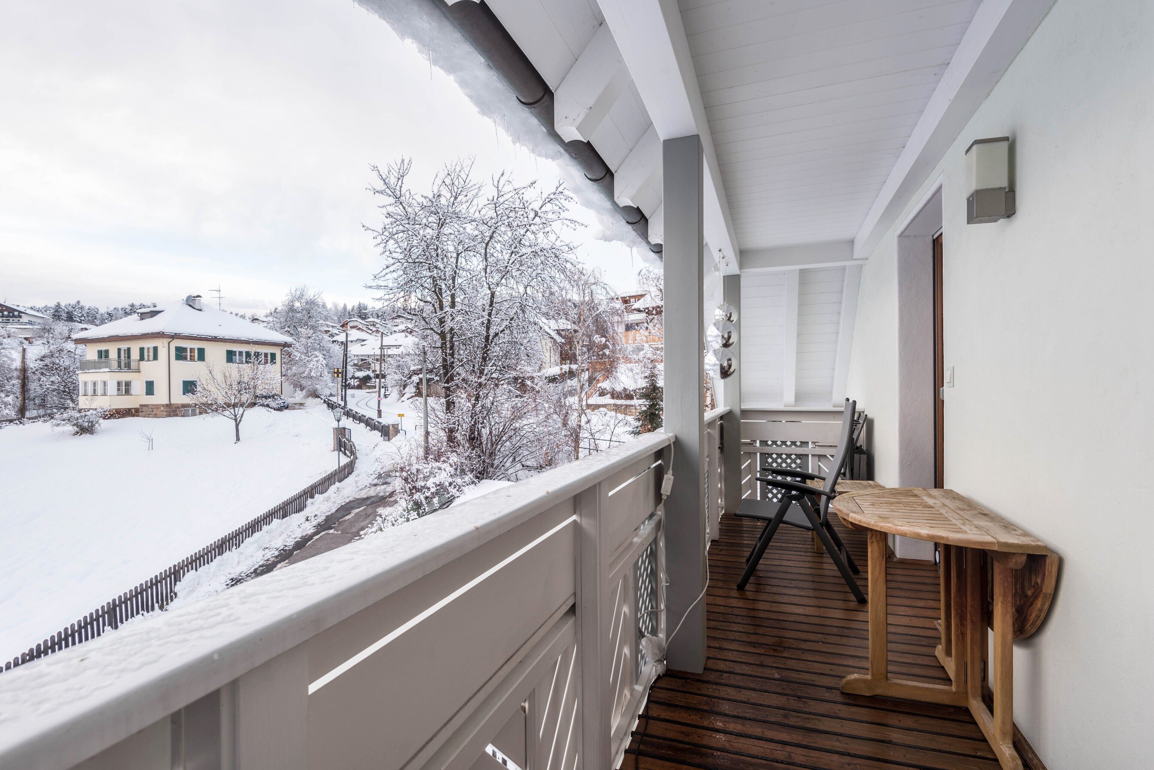A balcony with a wooden table and chairs, a white railing, and a view of a snow-covered landscape.
