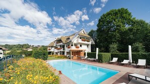 Een zwembad met een houten terras, ligbedden en een groot huis met balkons.