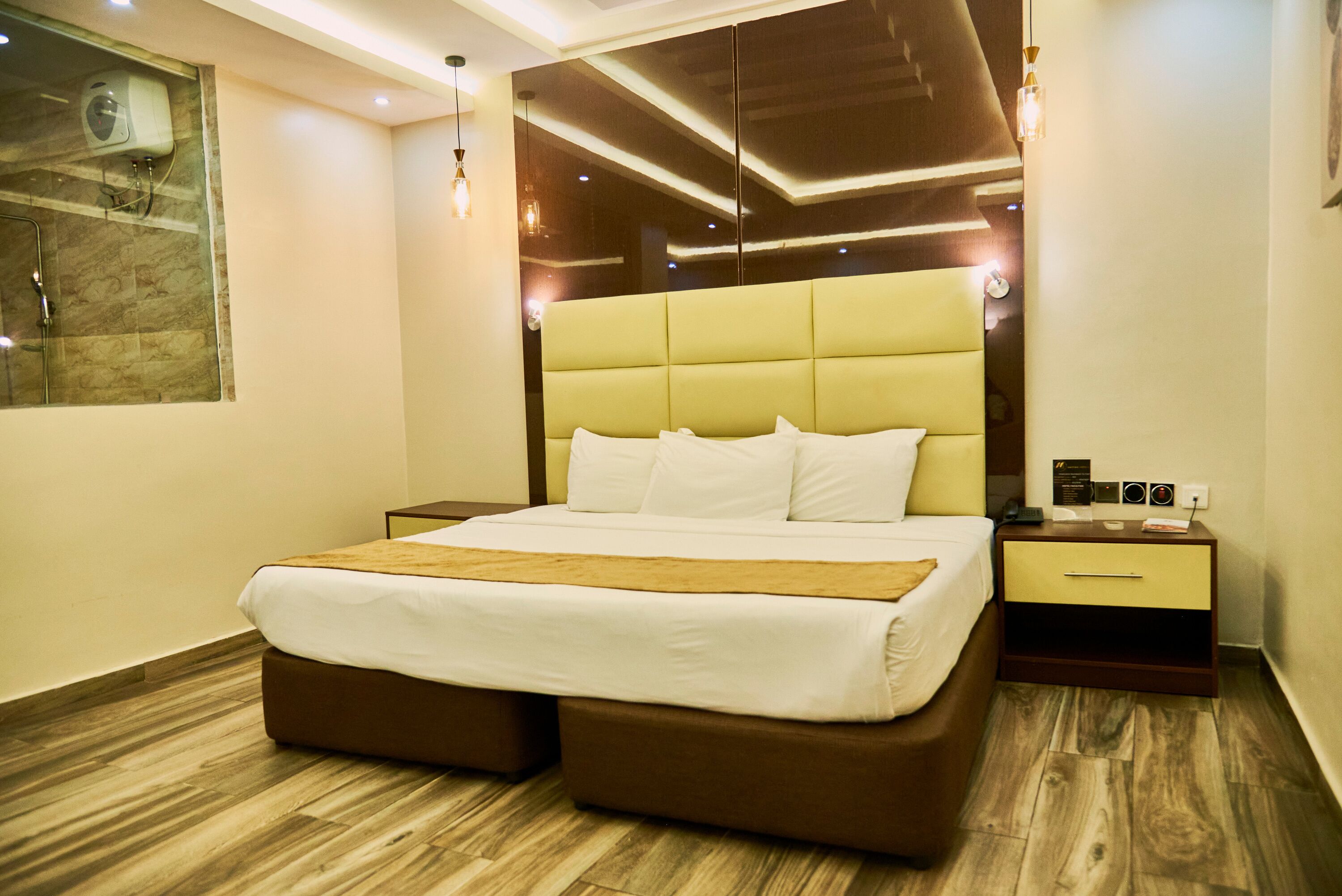 A modern hotel room with a large bed, wooden flooring, a nightstand with a lamp, and a wall-mounted air conditioning unit.