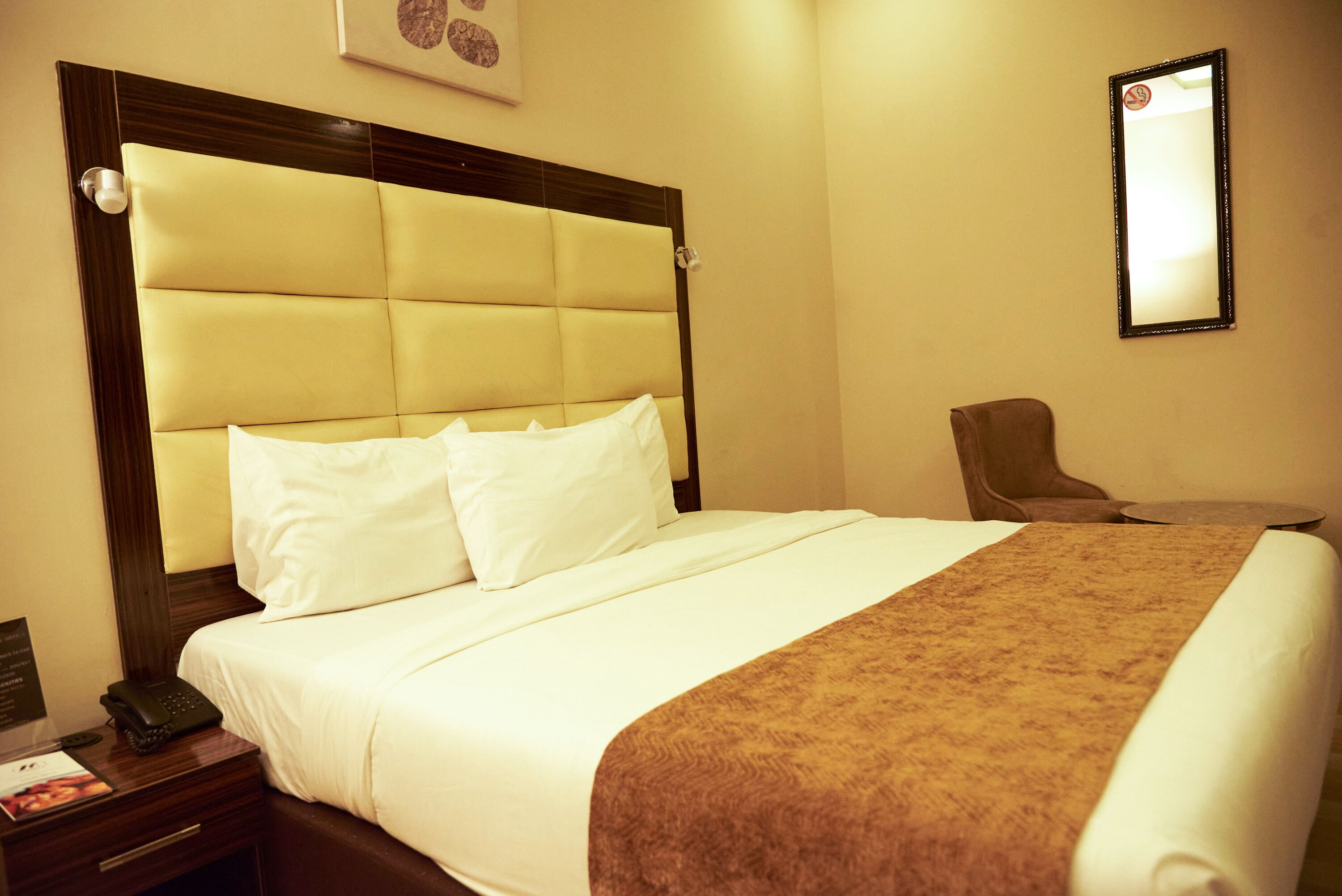Una habitación de hotel con una cama grande, una mesita de noche con un teléfono y una revista, una silla y un espejo en la pared.