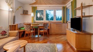A cozy living room with a sofa, dining table, chairs, a TV, and a kitchen area with a window. - Charming Apartment Tundra in Villa Anina with Pool (Klobenstein / Ritten)