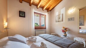 A hotel room with two beds, a window with curtains, a radiator, a bedside table with a vase of flowers, and a mirror on the wall.