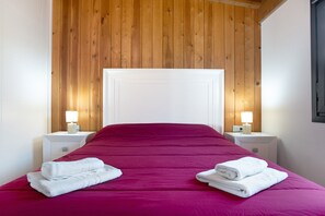 Een kamer met een houten plafond, een wit hoofdeinde, een rode spreidekker, twee witte nachtkastjes met lampen, en opgevouwen witte handdoeken op het bed.