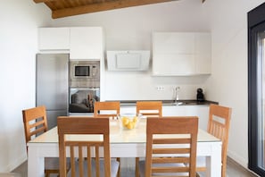 Una cocina moderna con armarios blancos, un refrigerador de acero inoxidable, un horno y un microondas. Hay una mesa de comedor con sillas de madera y un tazón de fruta.