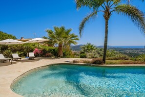 Piscina com espreguiçadeiras e guarda-sóis, cercada por palmeiras e vegetação, com vista para colinas ao longe.