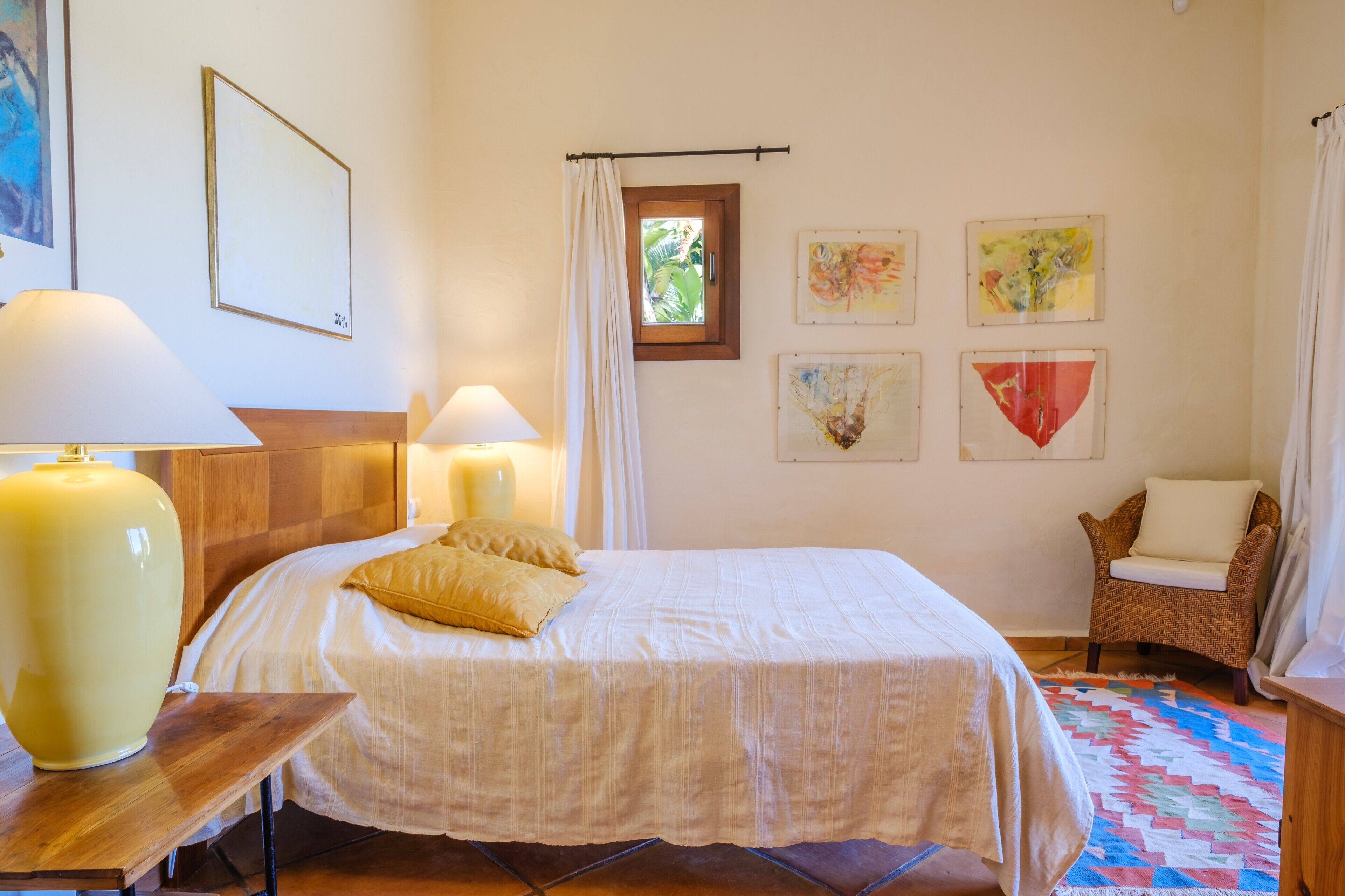Un dormitorio con una cama, dos lámparas de noche, un sillón de mimbre y decoración en la pared.