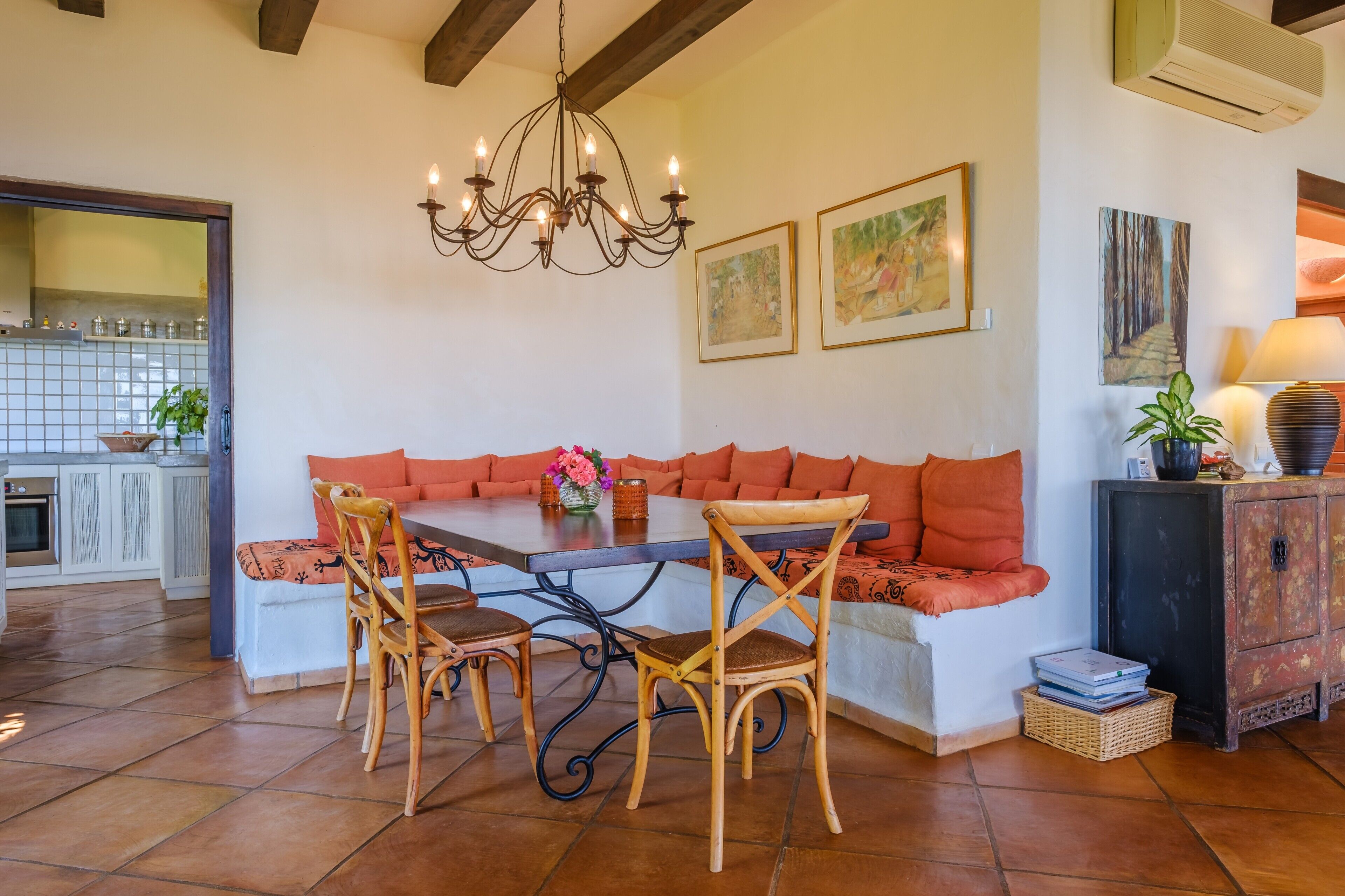 A dining area with a bench, chairs, a chandelier, and a kitchen in the background.