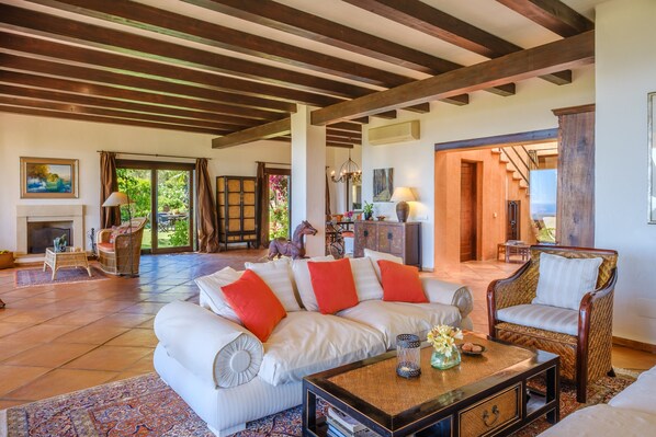 A spacious living room with a fireplace, a sofa with red cushions, a wicker chair, and a coffee table.