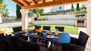 Une terrasse extérieure avec une table dressée pour le petit-déjeuner, des chaises en osier et une pergola au-dessus.