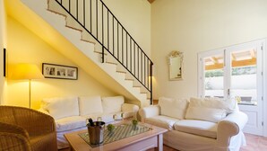 Un salon avec un escalier, deux canapés blancs, un fauteuil en osier, une table basse avec une bouteille et un verre, et une fenêtre donnant sur un paysage.