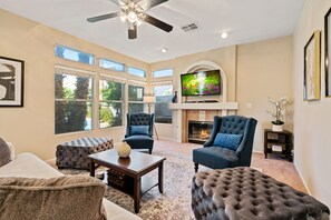 A spacious living room with a fireplace, large windows, and a ceiling fan.