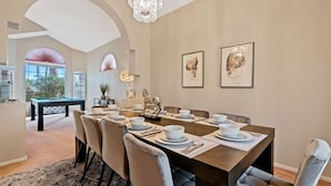 Uma sala de jantar com uma mesa comprida posta para seis pessoas, rodeada por seis cadeiras. Há uma mesa de bilhar ao fundo, e o cômodo possui um teto alto com um lustre.