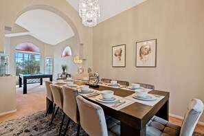 A dining room with a long table set for six, surrounded by six chairs. There is a pool table in the background, and the room has a high ceiling with a chandelier.