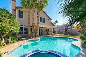 A swimming pool with a tiled edge, surrounded by palm trees and a house with a patio.