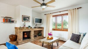 A living room with a fireplace, a television, a ceiling fan, and a window with curtains. - Rural Charm with Pool – Villa Damián 12 (Alcúdia)