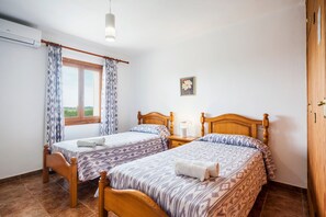 Una habitación con dos camas de madera, una ventana con cortinas, una mesita de noche con lámpara y un aire acondicionado de pared.