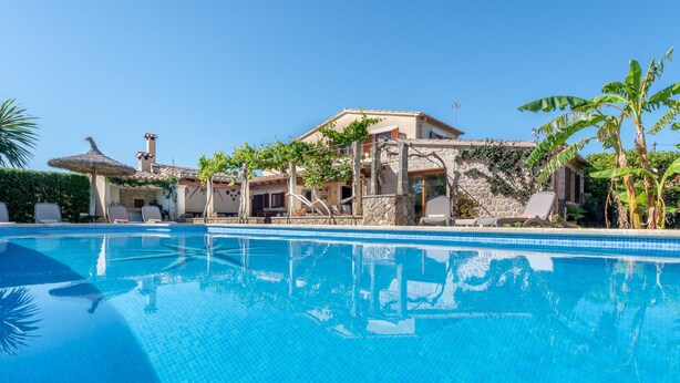 Ein Swimmingpool mit Liegestühlen, ein Steinhaus mit Ziegeldach und Palmen.