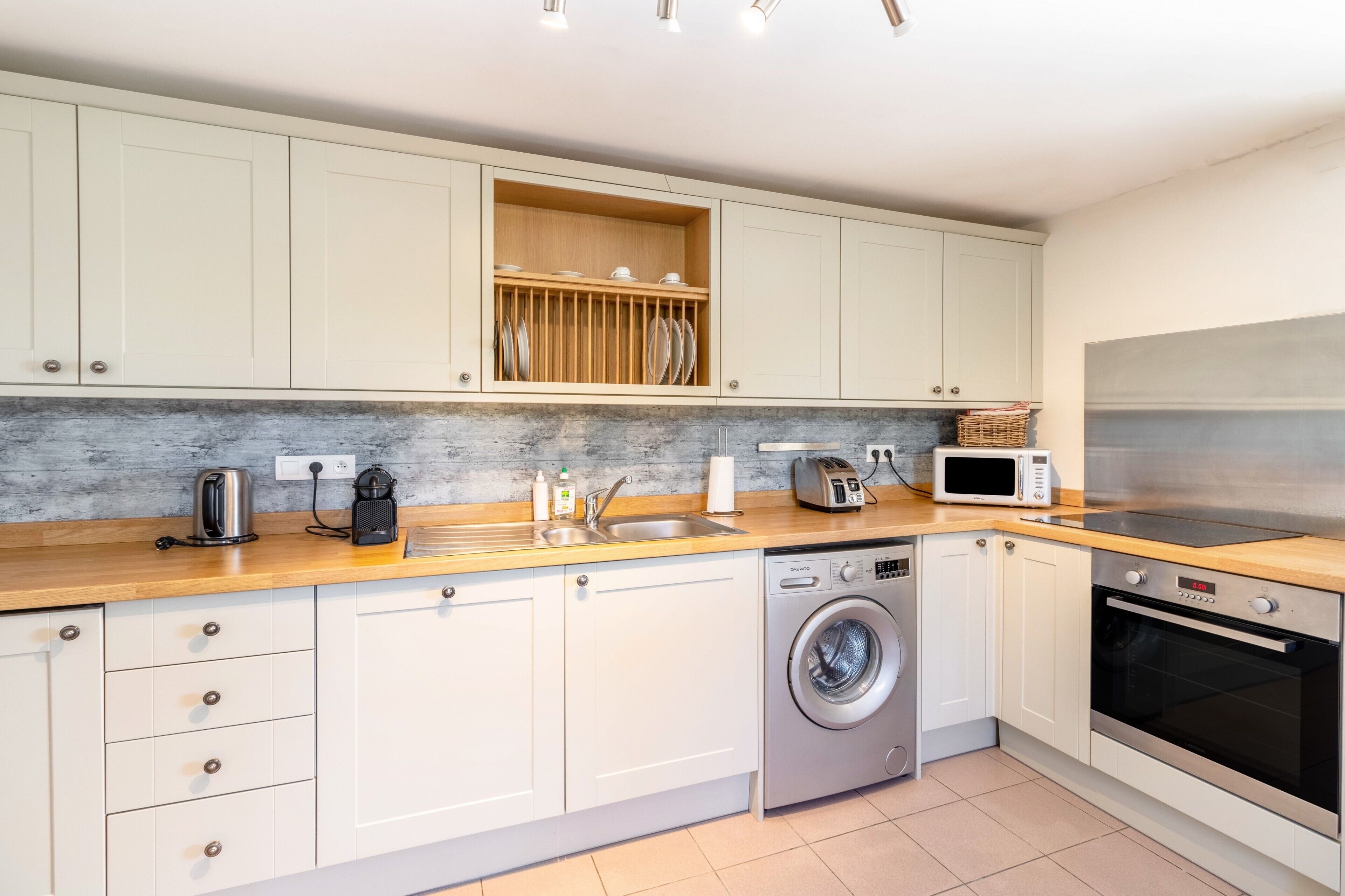 A compact kitchen with a washing machine, microwave, toaster, and a wooden shelf above the sink.