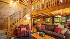 Interior de una cabaña de madera con una escalera, una sala de estar con sofá y sillón, y un área de cocina.