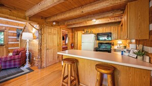 Interior de una cabaña de madera con una zona de cocina que incluye un refrigerador, microondas y taburetes de bar de madera.