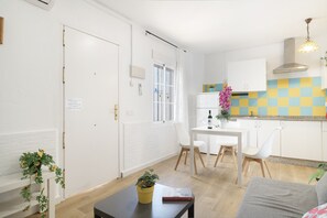 Moderni keittiö, jossa on valkoiset kaapit, ruokailutila pöytineen ja tuoleineen sekä olohuone, jossa on sohva.