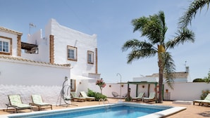 A pool area with lounge chairs, a palm tree, and a white building with windows.