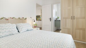 A bedroom with a bed, a wardrobe, a bedside table with a lamp, and a view into the kitchen.