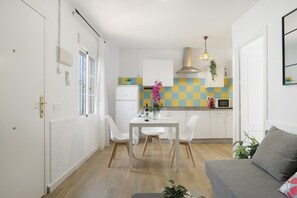 Een kleine keuken met witte kastjes, een eettafel met stoelen en een grijze bank.