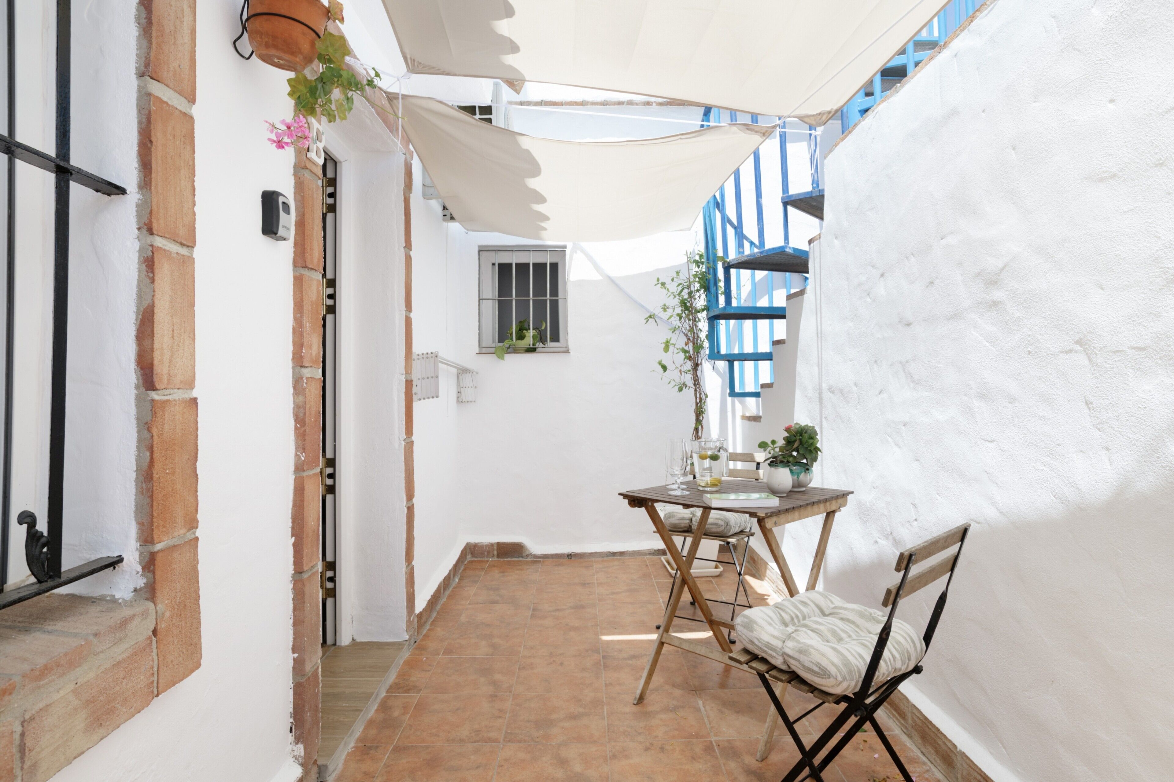A narrow outdoor patio with a table and chairs, a potted plant, and a white wall with blue accents.