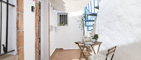 A narrow outdoor patio with a table and chairs, a potted plant, and a white wall with blue accents.