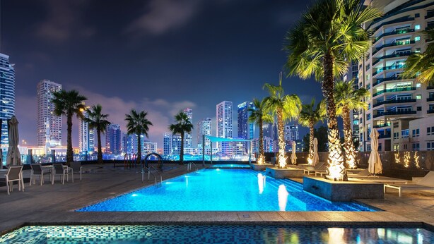 Uma piscina à noite com um horizonte urbano ao fundo, cercada por palmeiras e espreguiçadeiras.