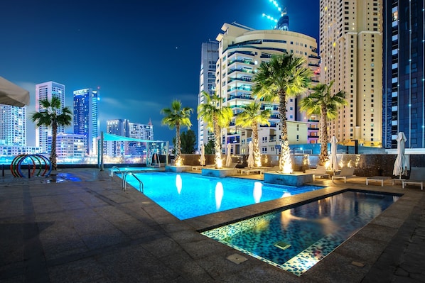 A rooftop pool area with palm trees, lounge chairs, and a view of the city skyline at night.