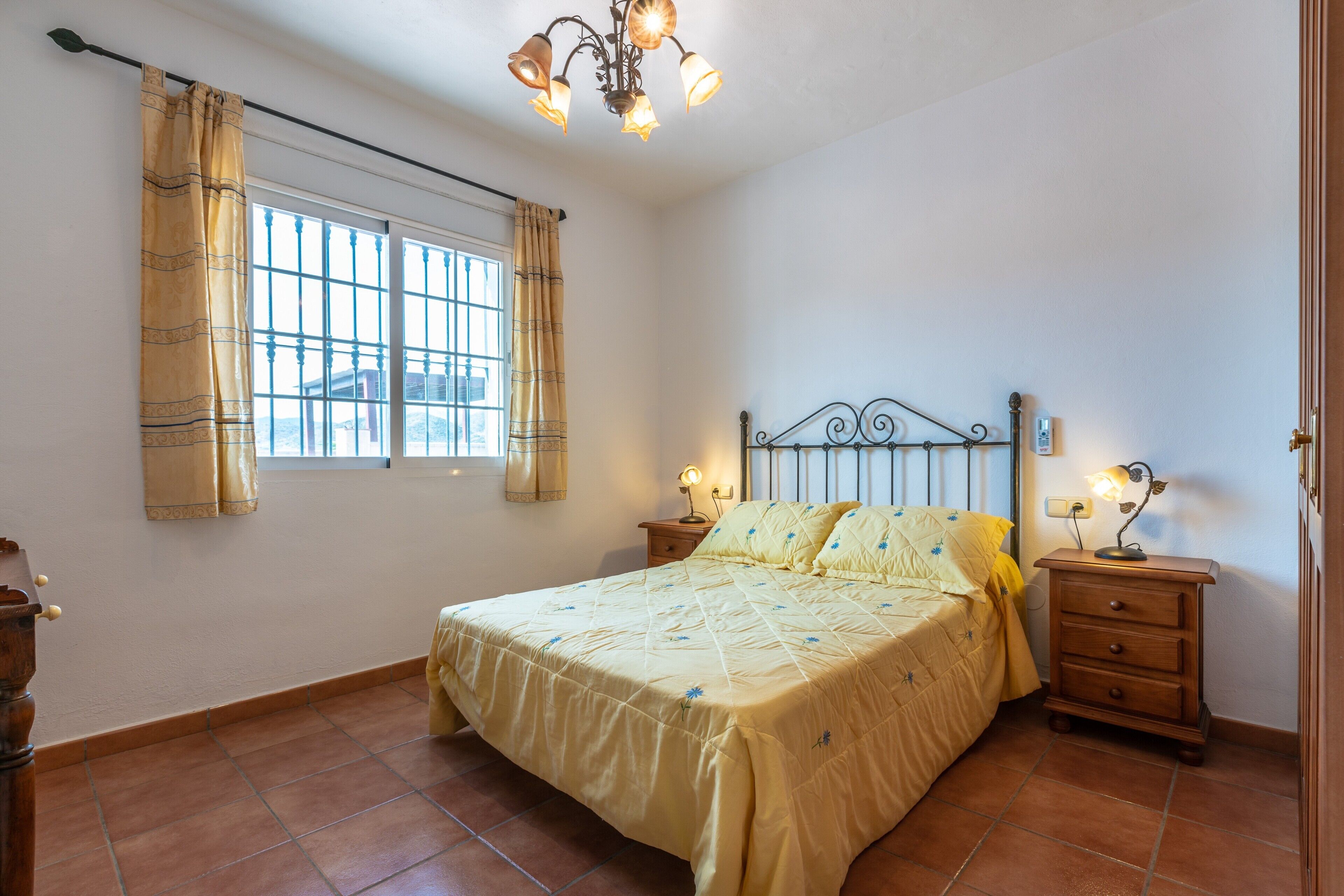 Un dormitorio con cama, dos mesitas de noche, un tocador, una ventana con cortinas y una araña.