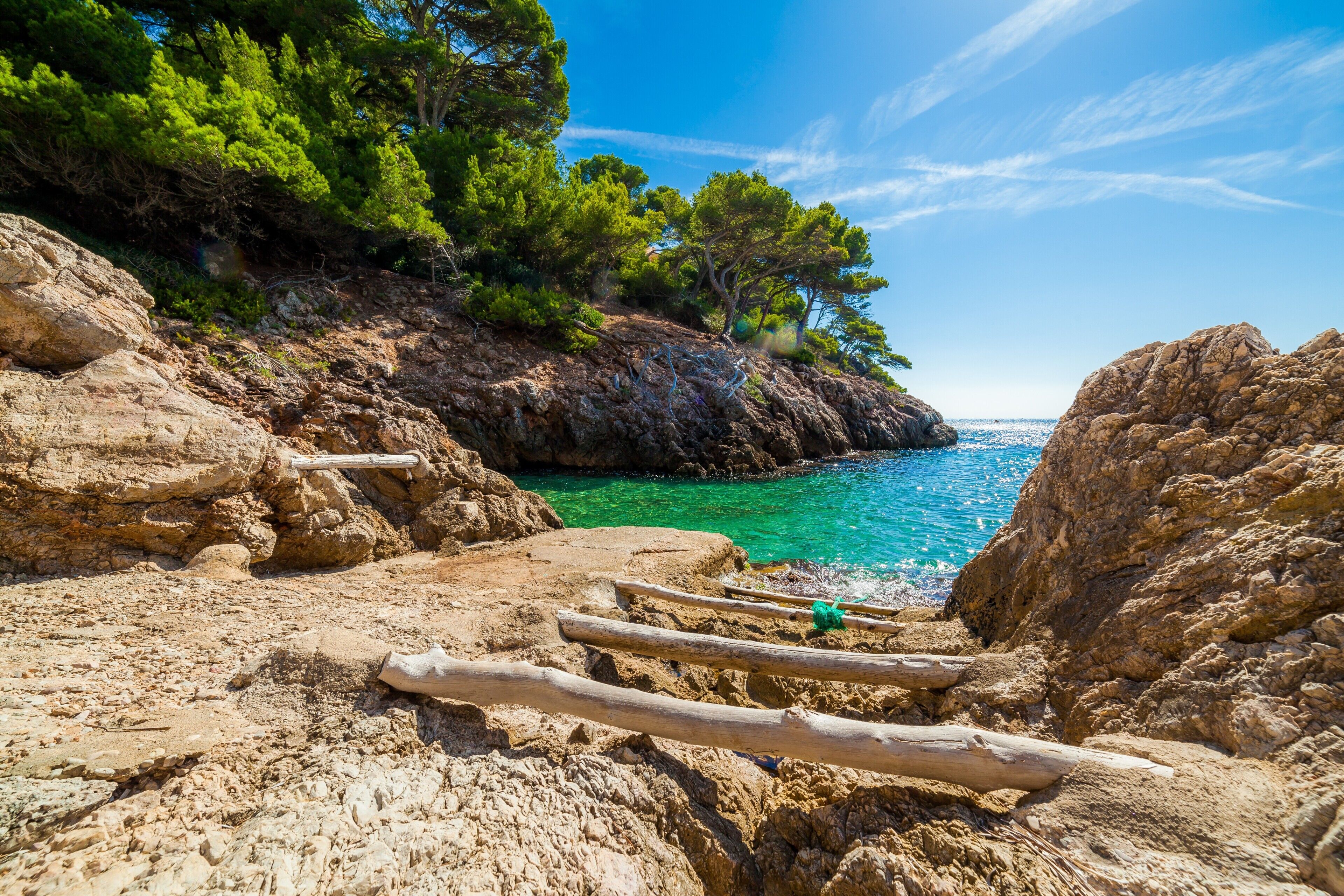 Paesaggio costiero roccioso con acque turchesi cristalline, alberi verdi rigogliosi e un sentiero di legno che conduce verso il mare.