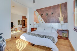A modern hotel room with a large bed, a wall-mounted artwork, a desk with a chair, and a television.