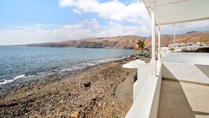 Un hôtel situé sur la côte, avec une plage rocheuse, des murs extérieurs blancs et une vue sur la mer et les collines.