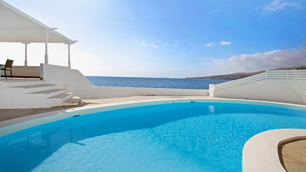 Une piscine avec vue sur la mer, un espace salon couvert et un bâtiment blanc.