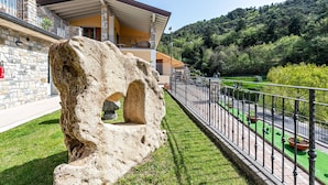 Ein Steinhaus mit einem Rundbogen, einem Balkon mit Geländer und Blick auf eine grüne Landschaft mit Bäumen und einer Hügelkulisse im Hintergrund.
