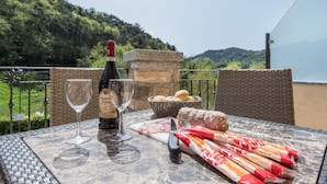 Ein Tisch mit einer Weinflasche, Gläsern, Brot und einem Korb mit Brötchen auf einem Balkon mit Blick auf einen bewaldeten Hügel.
